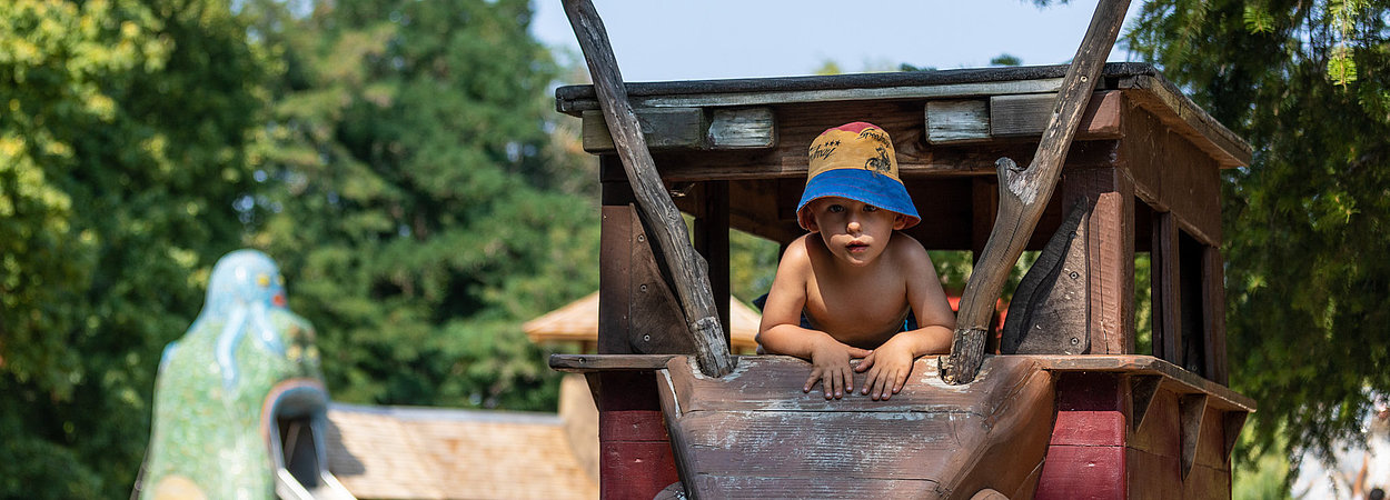 Ein kleiner Junge guckt aus einem Spielgerät auf der Spiellandschaft am Lehmdorf heraus in die Kamera