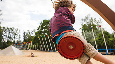 Ein Kind schwingt auf einer Schaukel, im Hintergrund ist eine Spiellandschaft zu sehen